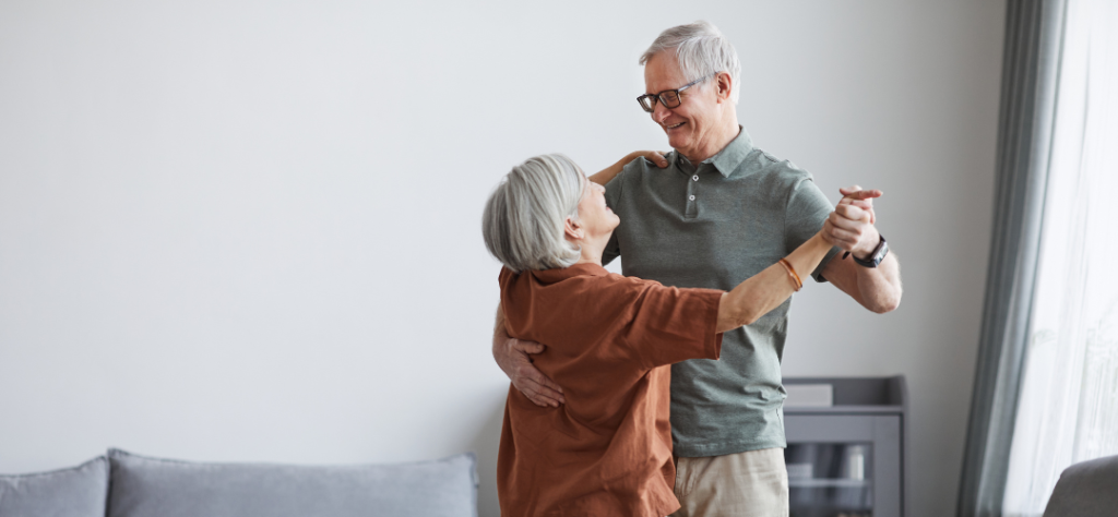 Casal de idosos dançando e sorrindo em casa, ilustrando a qualidade de vida e alegria no envelhecimento.