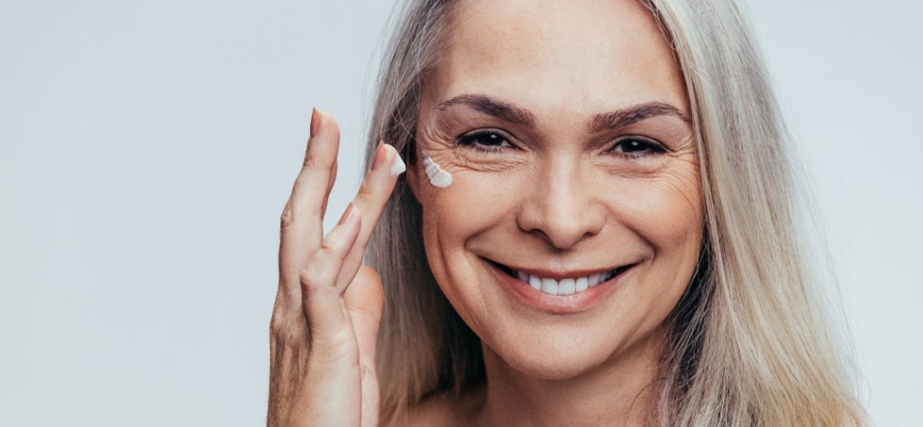 Mulher madura sorridente aplicando creme no rosto, representando cuidados com a pele e saúde no envelhecimento.