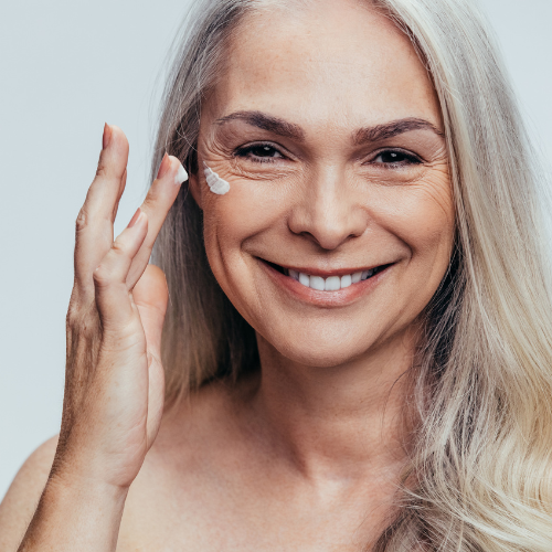 Longevidade, envelhecimento e a ciência de viver mais 28 Mulher madura sorridente aplicando creme no rosto, representando cuidados com a pele e saúde no envelhecimento.