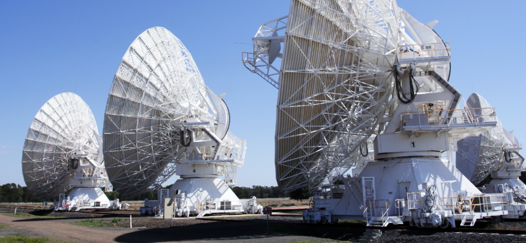 2026 - Telescópios espaciais: quando a humanidade decidiu enxergar além de si mesma 1 Os Equipamentos Estrutura: A imagem foca em três grandes antenas parabólicas (e parte de uma quarta ao fundo) pintadas de branco. Elas possuem uma estrutura complexa de treliças metálicas na parte traseira para suporte. Mobilidade: É possível notar que as bases dessas antenas estão montadas sobre trilhos ferroviários. Isso permite que os cientistas alterem a distância entre os telescópios, criando um interferômetro que funciona como um único telescópio gigante. Componentes: No centro das parábolas, há receptores sustentados por hastes, responsáveis por captar as ondas de rádio vindas do espaço.