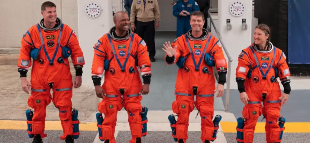 Os quatro astronautas da missão Artemis II caminhando com trajes espaciais laranjas da NASA.